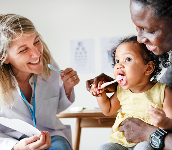 Mother holding her toddler while dentist shows toddler how to brush their teeth