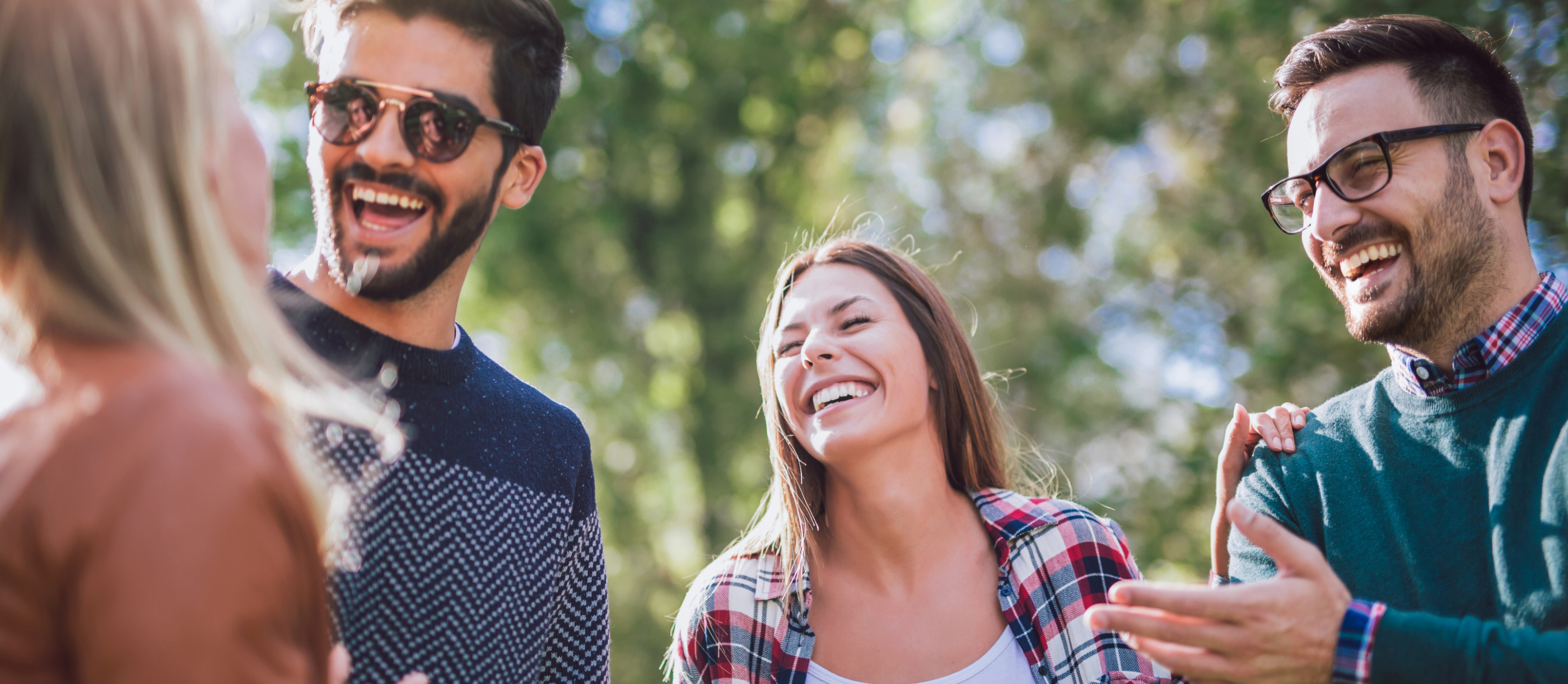 Four adults laughing together outdoors on a sunny day